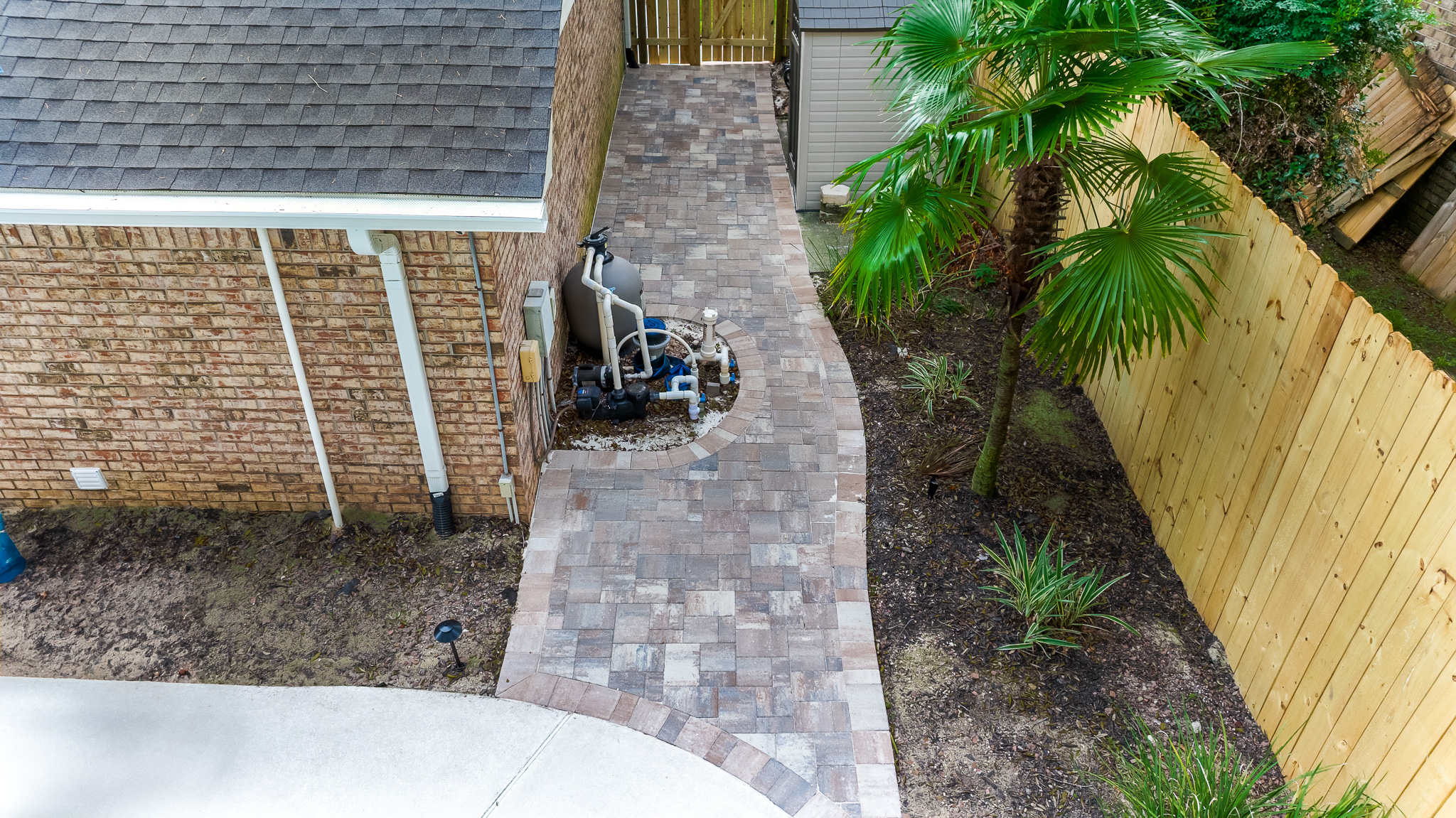 Paved walkway between brick house and wooden fence with pool equipment