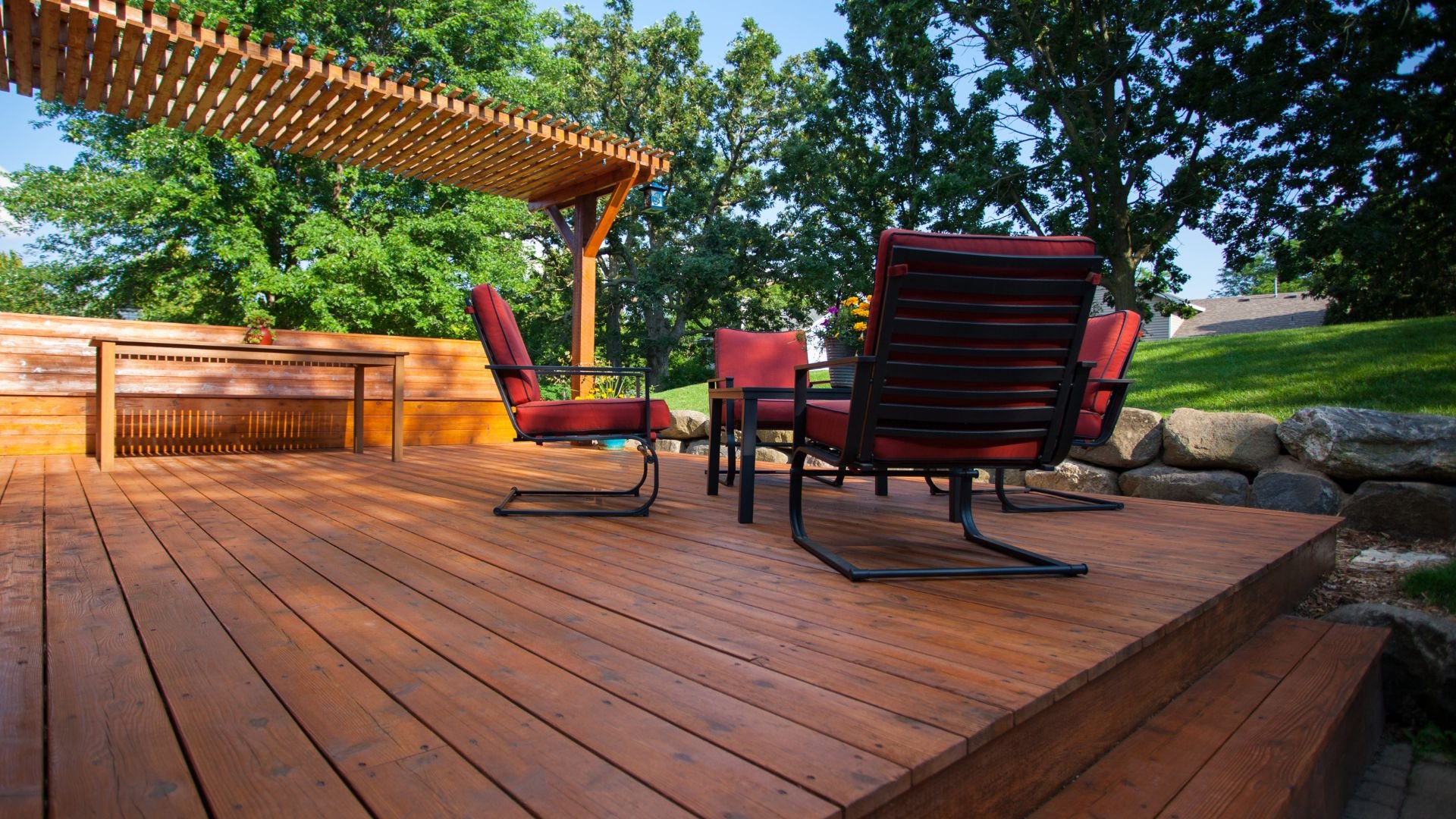 Wooden deck with red chairs, pergola, and stone border on sunny day